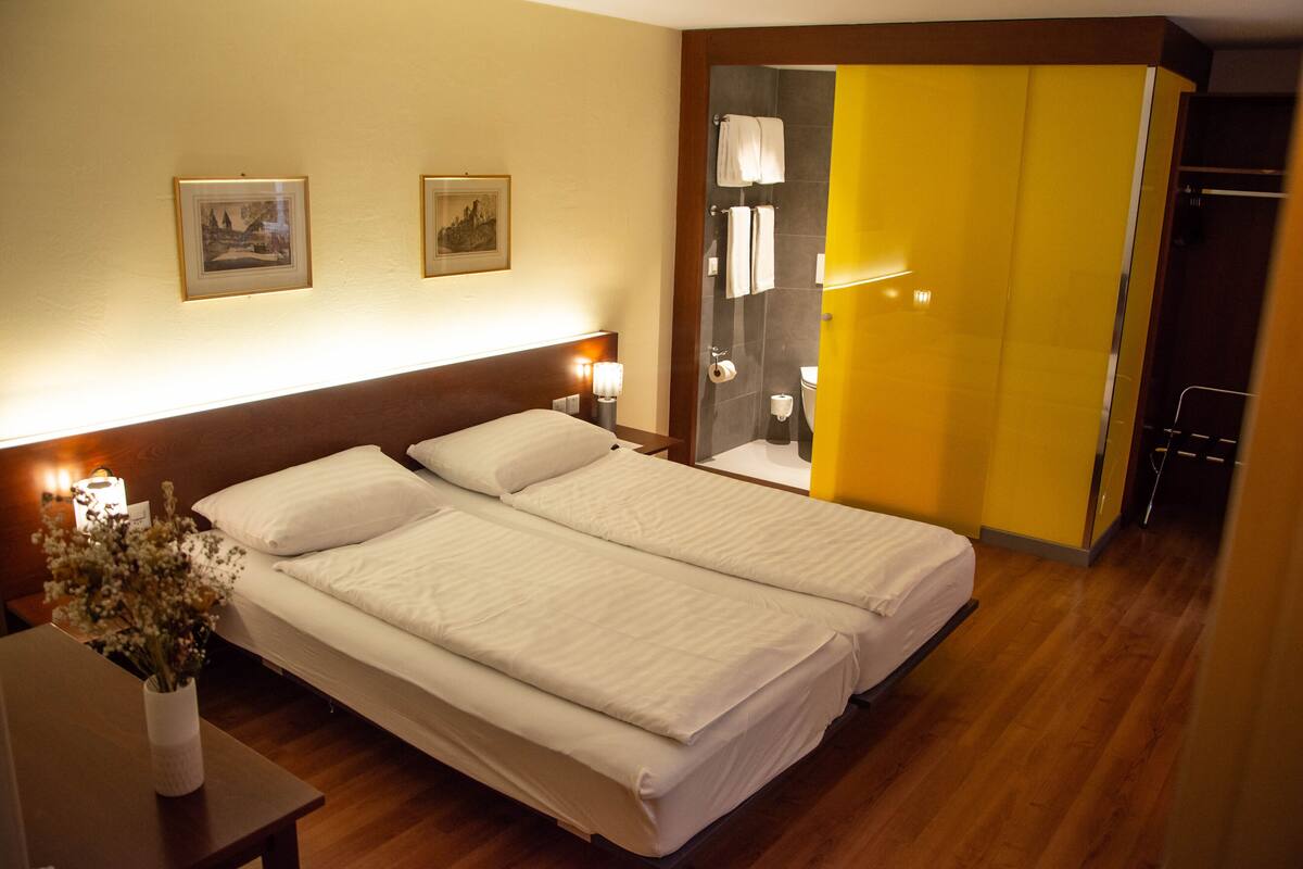 Habitació d'hotel amb dos llits individuals, un capçal de llit de fusta, quadres amb marc a la paret, una taula de nit amb un ram i un bany amb porta de vidre.