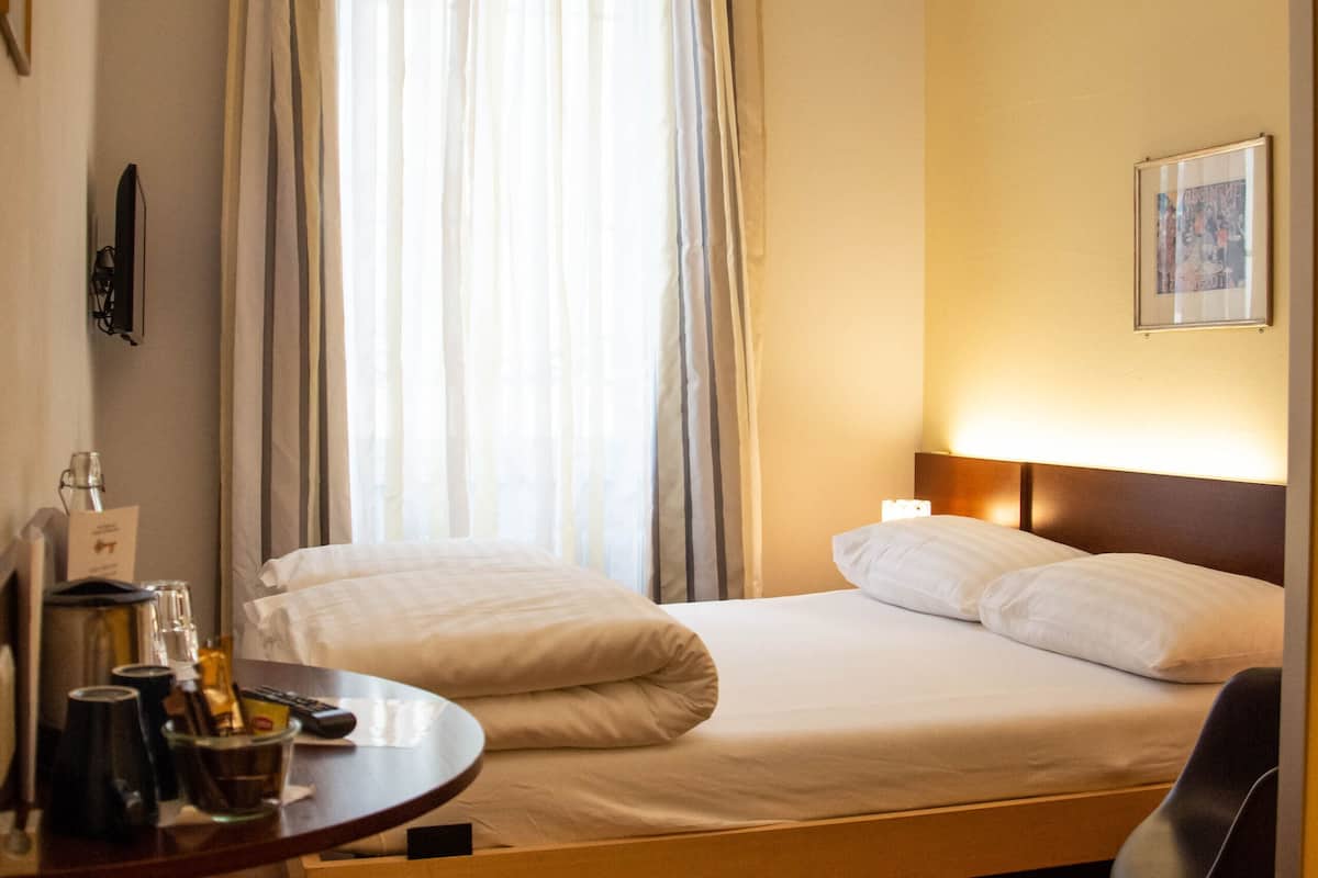 Una habitació d'hotel amb un llit, una taula de nit amb una làmpada, una cadira i una finestra amb cortines.