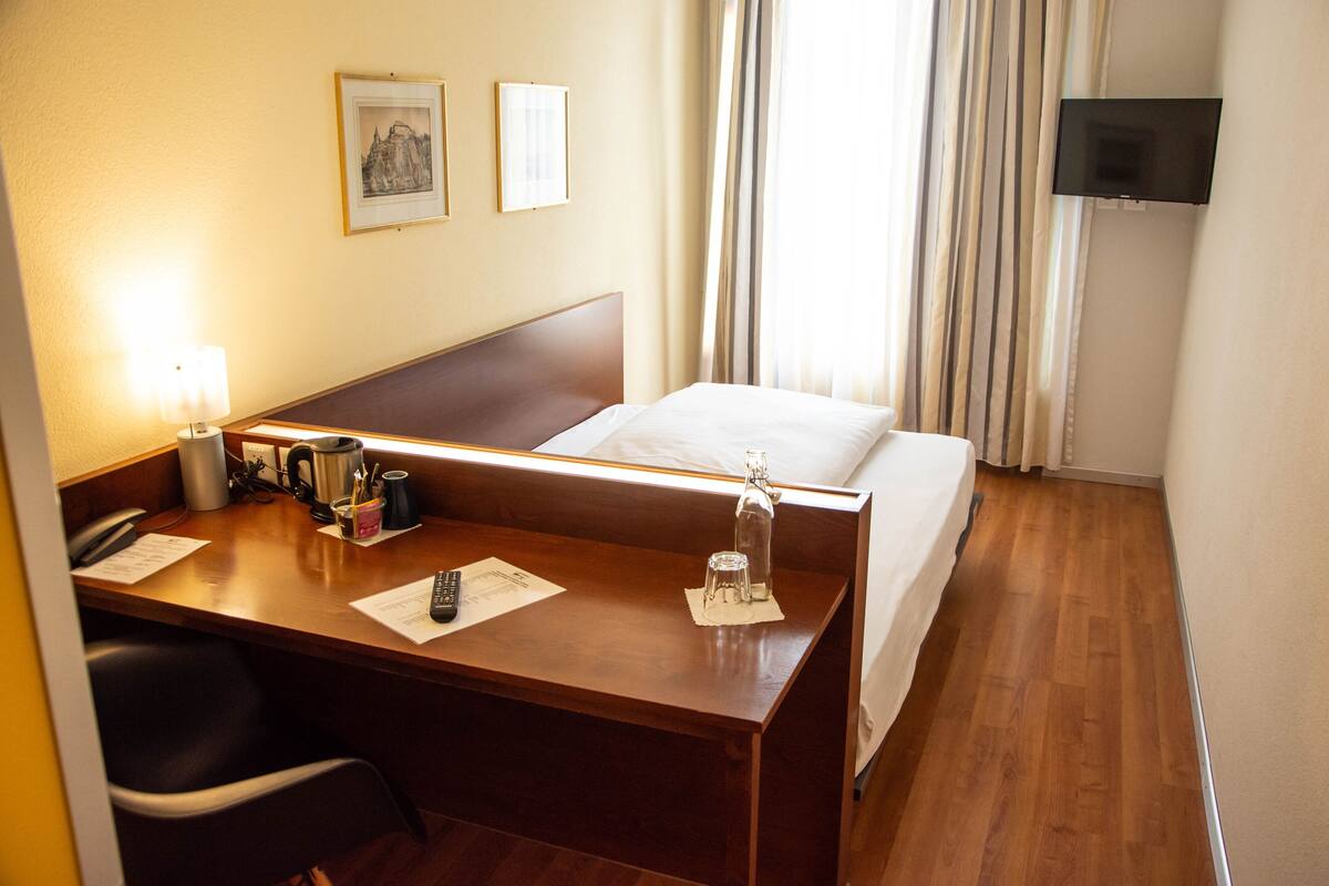 Habitació d'hotel amb escriptori de fusta, un llit, televisió penjada a la paret i una finestra amb cortines.