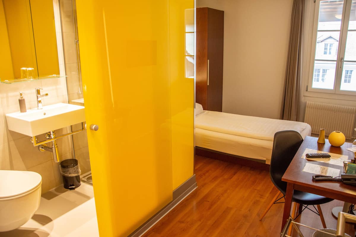 Una habitació d'hotel amb una paret d'accent groga, un llit, una taula de treball amb una cadira, una finestra amb vistes i un bany amb un lavabo i un vàter.