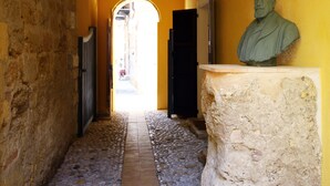 A narrow stone-paved alleyway with a statue on a pedestal, wooden beams on the ceiling, and a doorway leading to a brighter area. - Khalisah (Palermo)