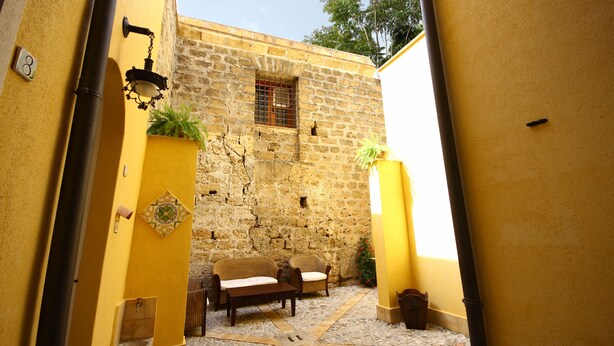 A narrow courtyard with a stone wall, a bench, and two wicker chairs. - Khalisah (Palermo)