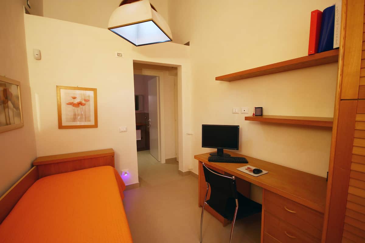 A bedroom with a bed, a desk with a computer, a chair, and a shelf with books.