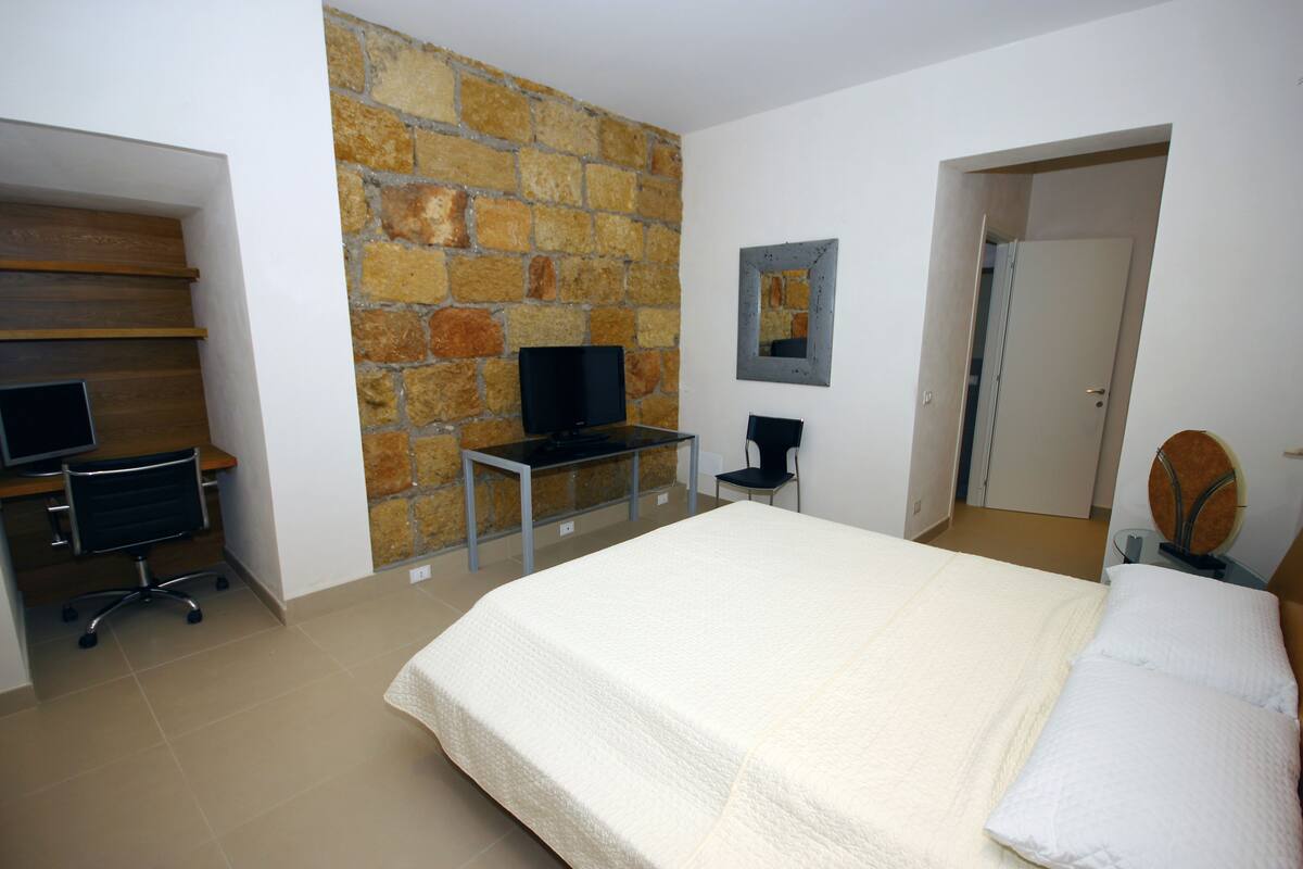 A bedroom with a large bed, a desk with a computer, a chair, and a stone wall.