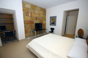 A bedroom with a large bed, a desk with a computer, a chair, and a stone wall.