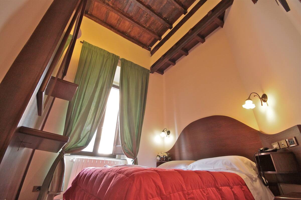 A bedroom with a wooden ceiling, a bed with a red blanket, a nightstand with a lamp, and a window with green curtains.