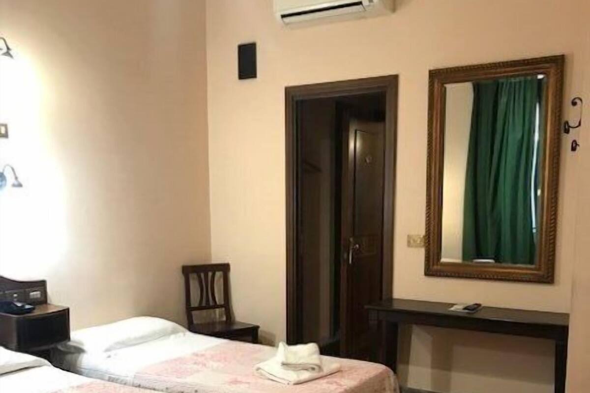 A hotel room with two beds, each with a floral bedspread and neatly folded towels. There is a wooden chair, a small table, and a mirror on the wall. An air conditioning unit is mounted on the wall above the door.