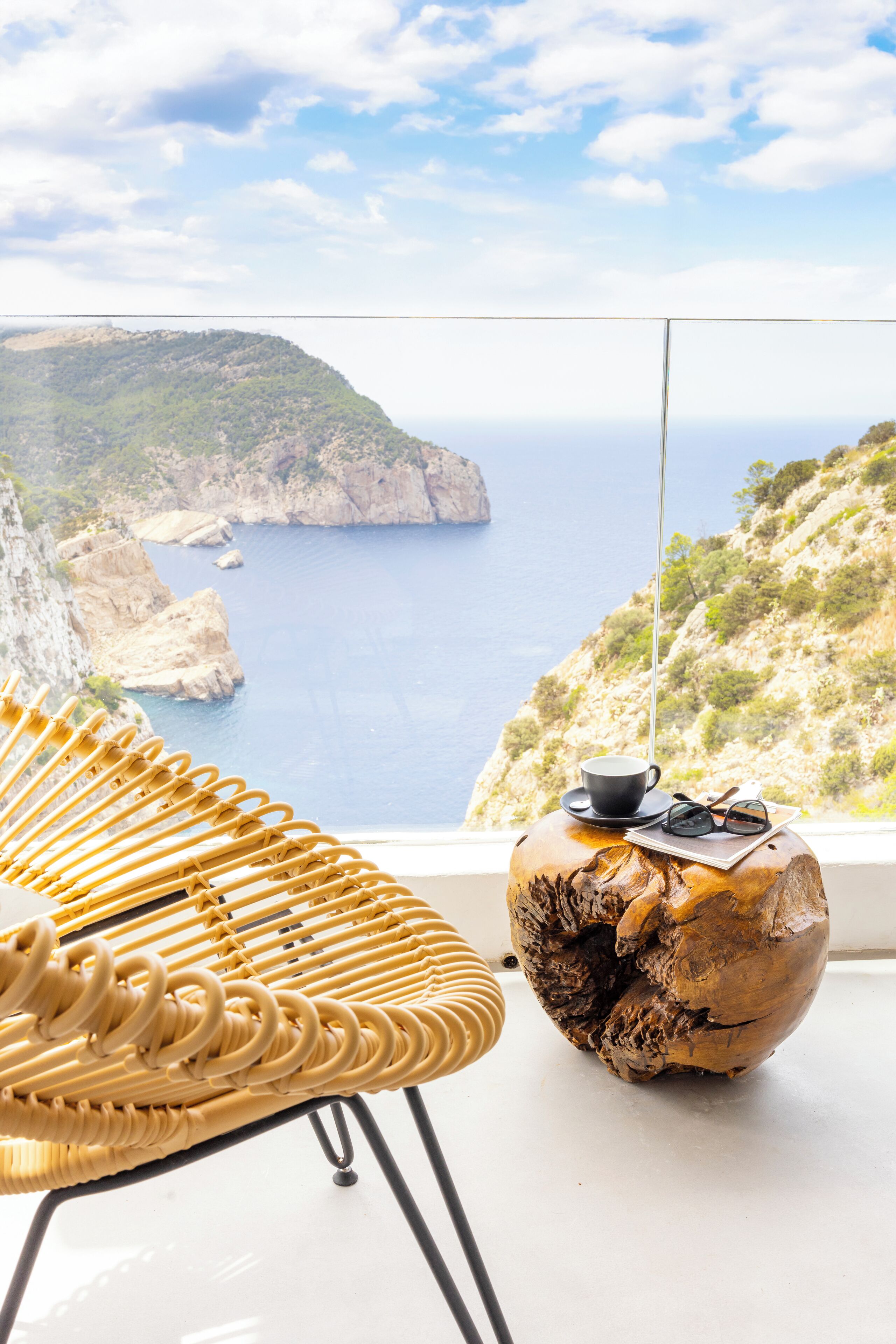 A wicker chair and a wooden table with a cup and sunglasses on it, overlooking a coastal landscape with cliffs and the sea.