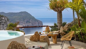 Zona junto a la piscina con vista al mar, una mesa de madera, una silla de mimbre y una roca grande.