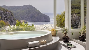 Een wellnessruimte met een jacuzzi, handdoeken en potplanten, met uitzicht op een kustlandschap met kliffen en de zee.