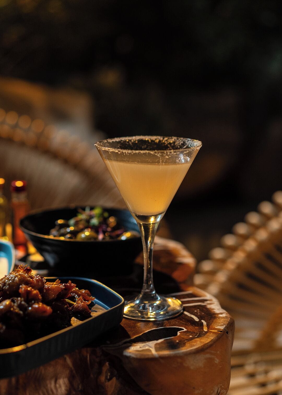 A cocktail in a martini glass with a salted rim, a bowl of food, and a bottle in the background.