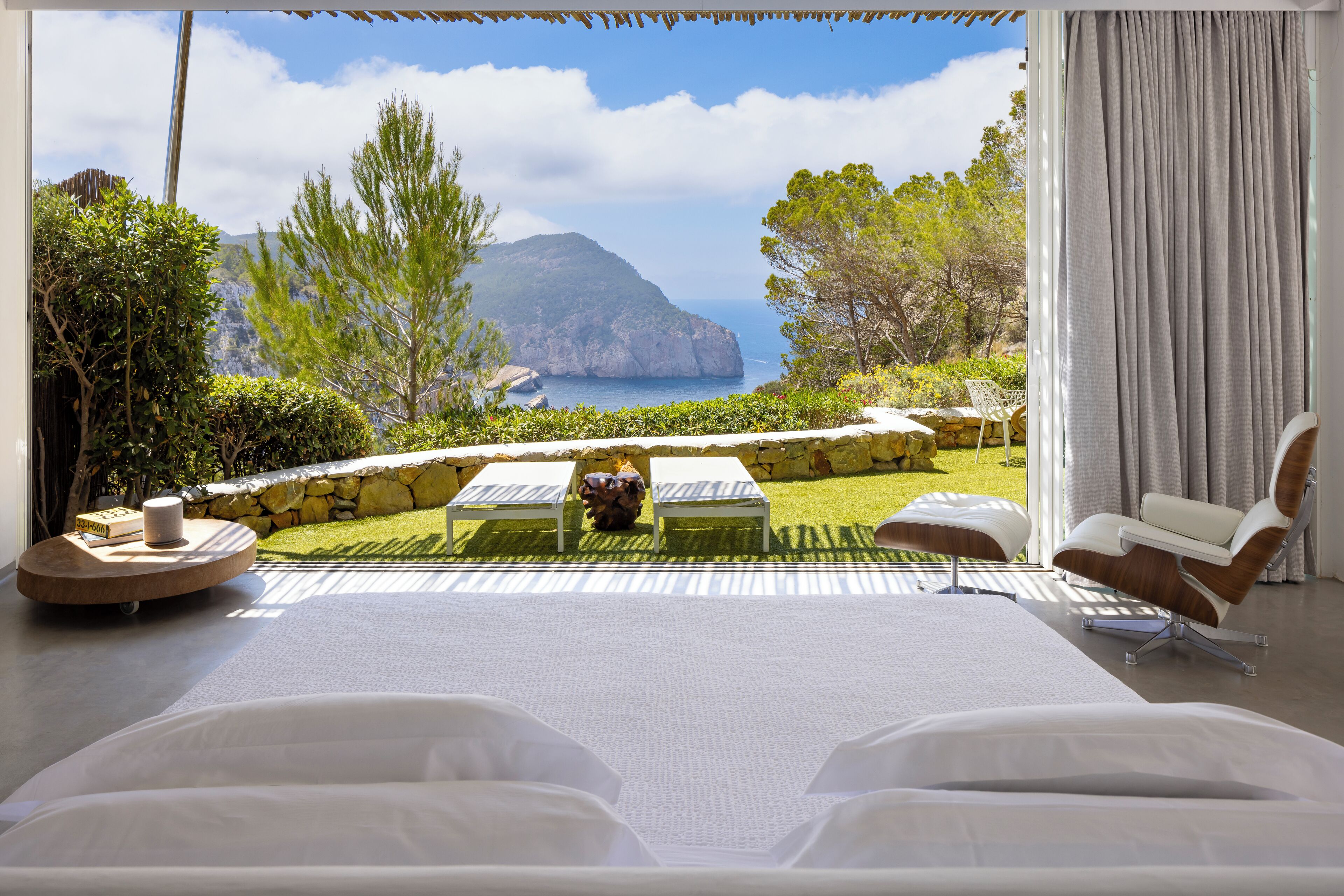 Ein Schlafzimmer mit einem großen Bett, zwei Sessel, einem kleinen Tisch und Blick auf das Meer und die Berge.