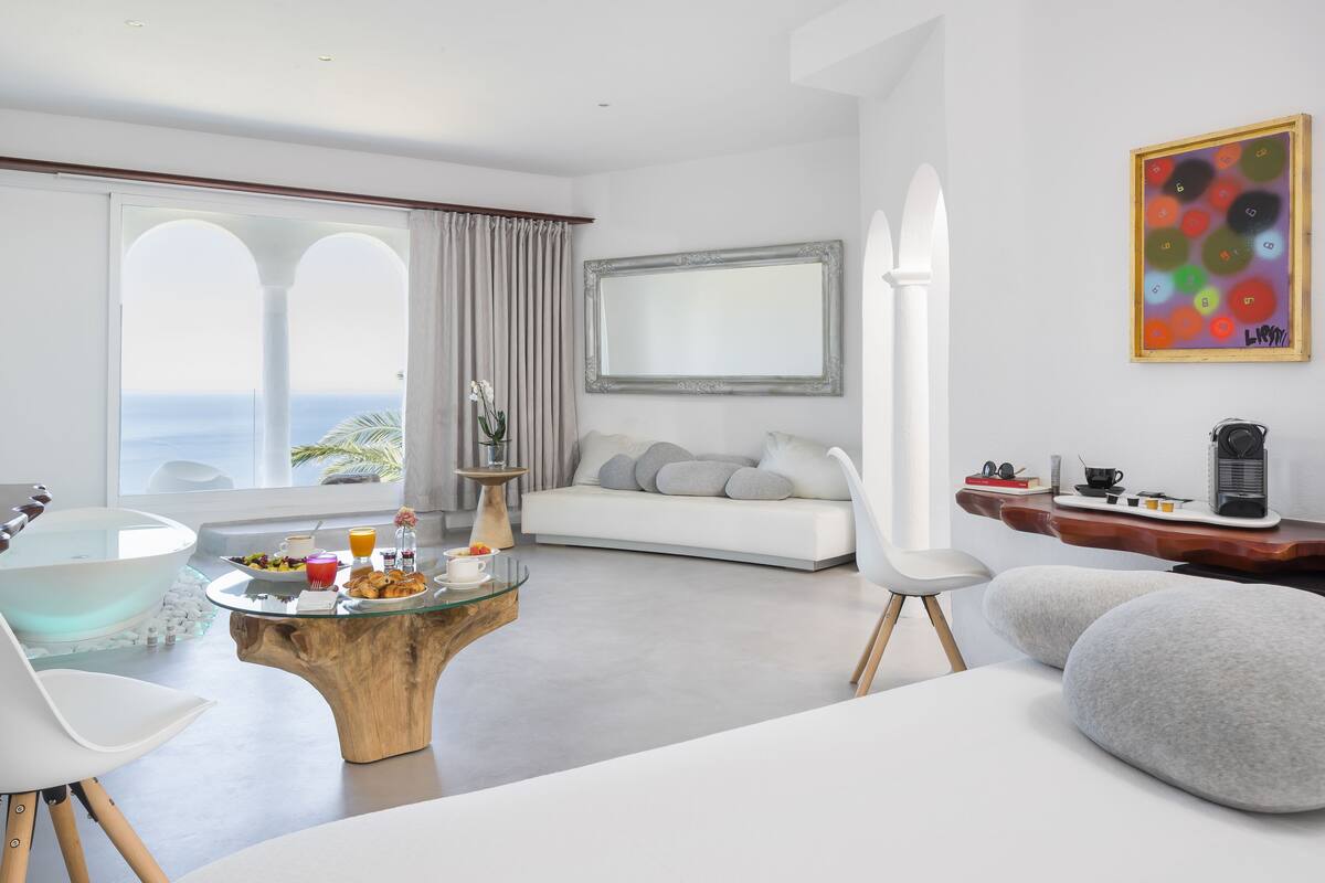 Ein modernes Wohnzimmer mit einem Glastisch, weißen Sofas, einem Holztisch und einem großen Fenster mit Meerblick.