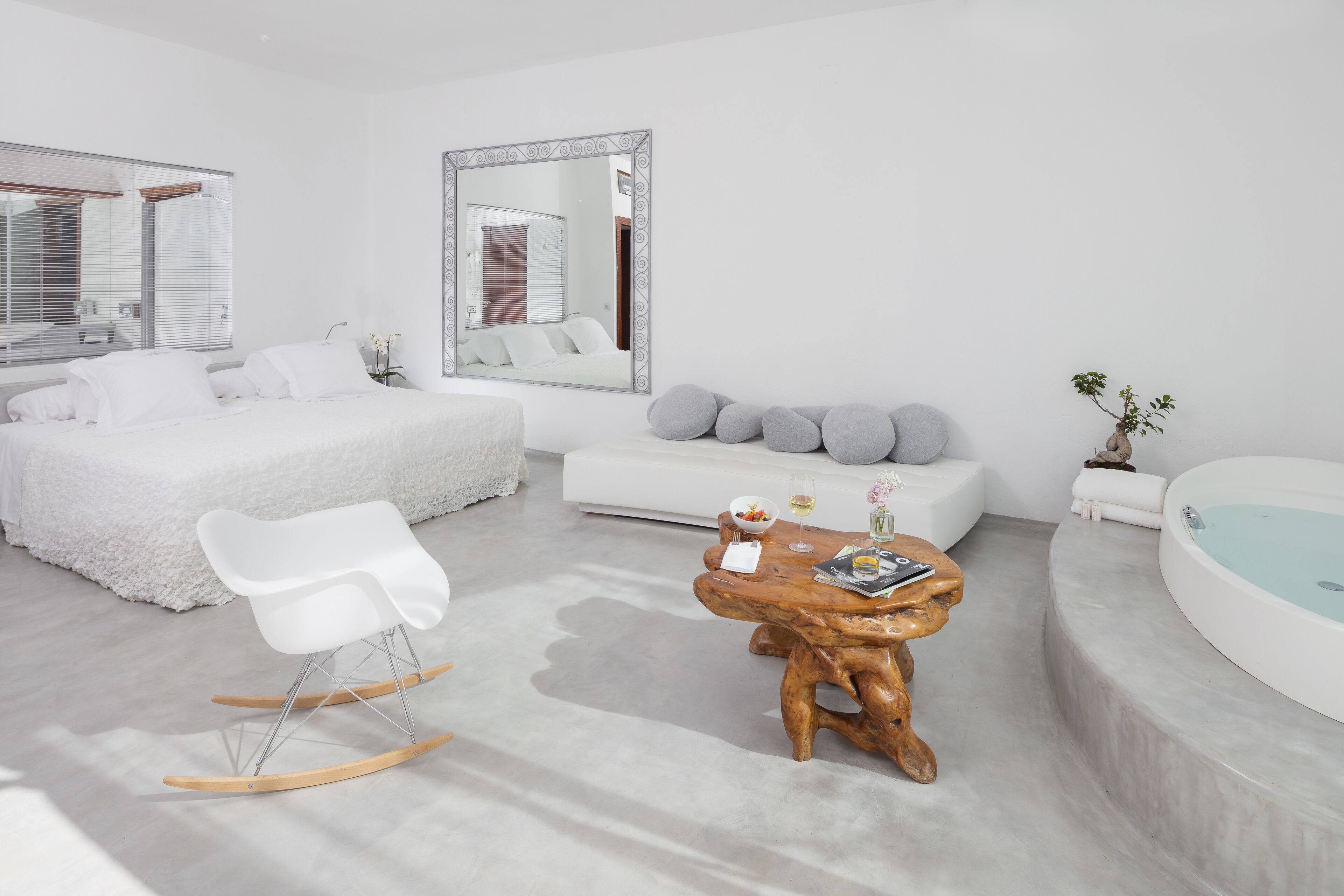 Un dormitorio moderno y minimalista con una cama grande, una mecedora blanca, una mesa de centro de madera, un espejo grande, un sofá blanco y una bañera.