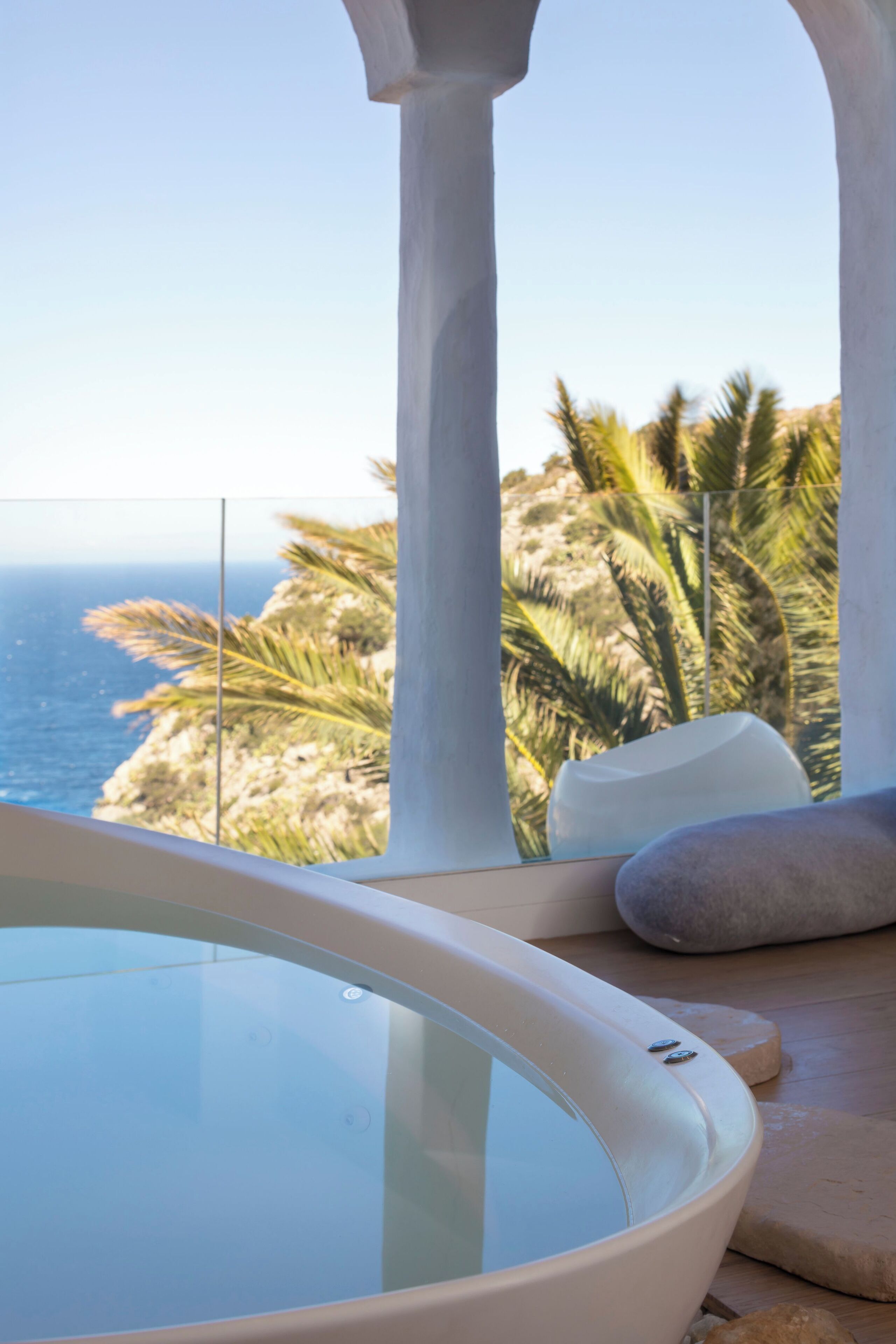 Un área de spa con un jacuzzi de paredes de vidrio, una zona de estar de piedra y vistas al océano y palmeras.