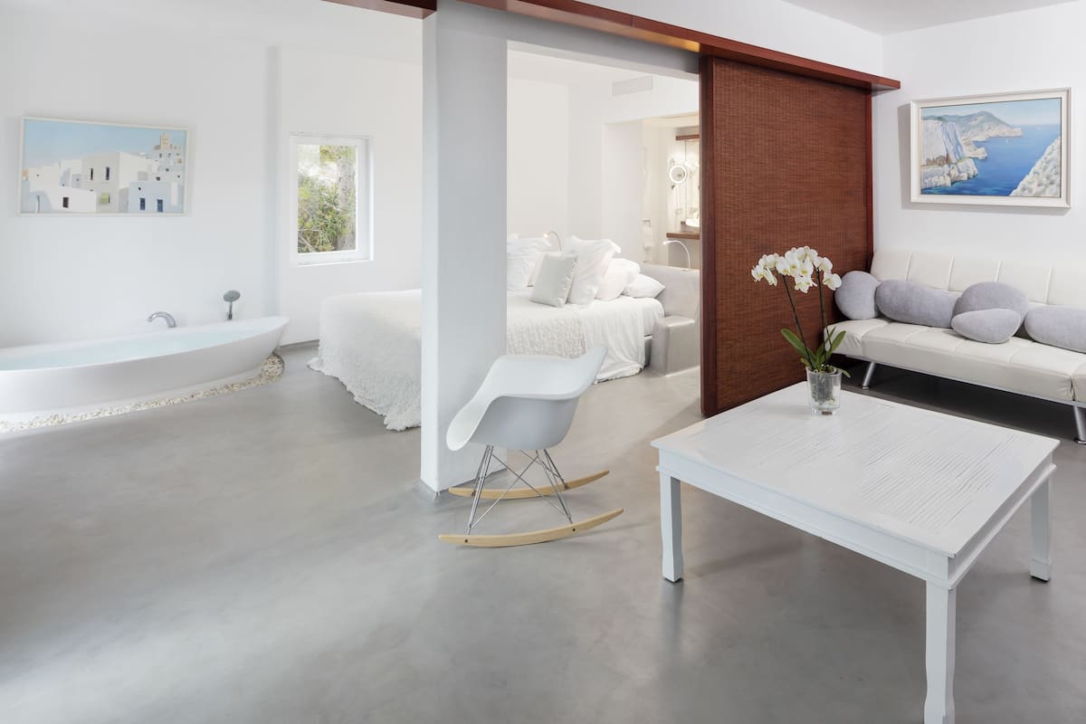 Ein modernes, minimalistisches Schlafzimmer mit einem großen Bett, einem weißen Schaukelstuhl, einem weißen Tisch und Meerblick durch das Fenster.