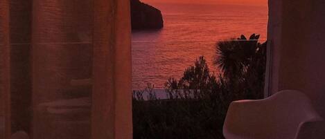 A room with a view of the sea at sunset, a white chair, and a small round table.