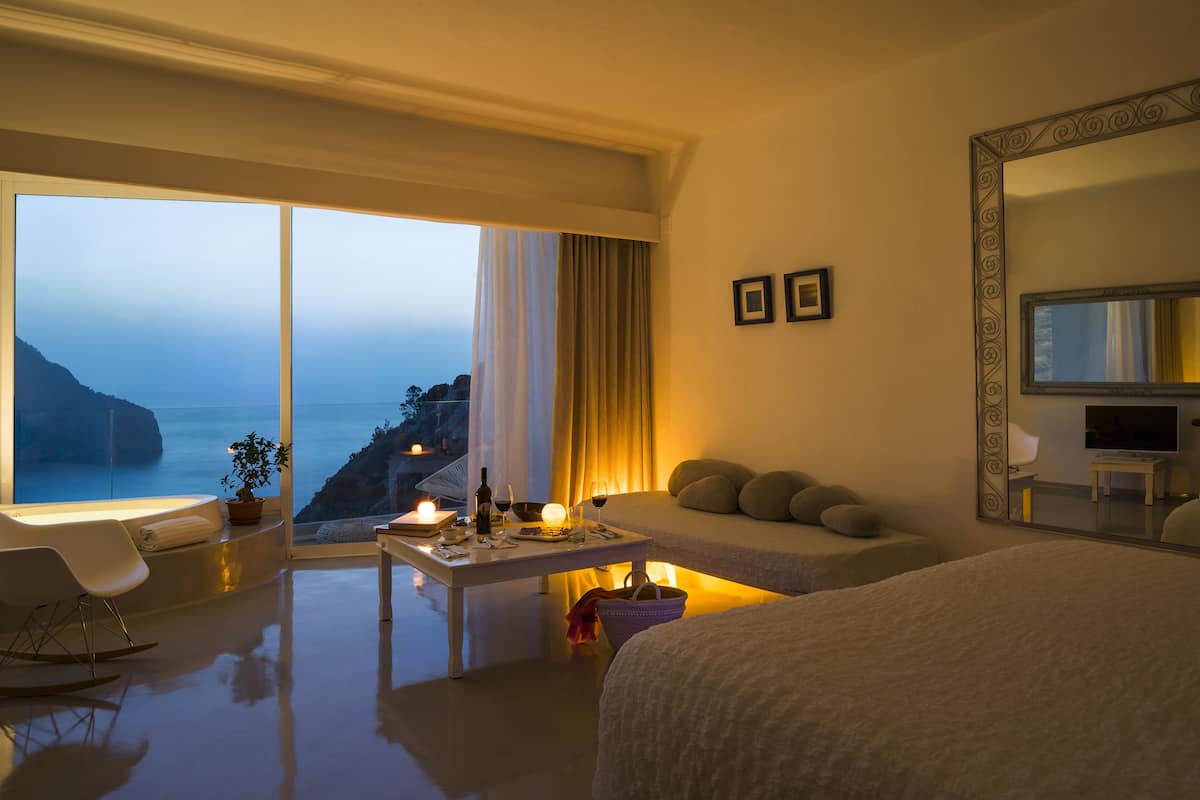 Ein modernes Hotelzimmer mit einem großen Bett, einer Couch, einem Esstisch mit Kerzen und Blick aufs Meer.