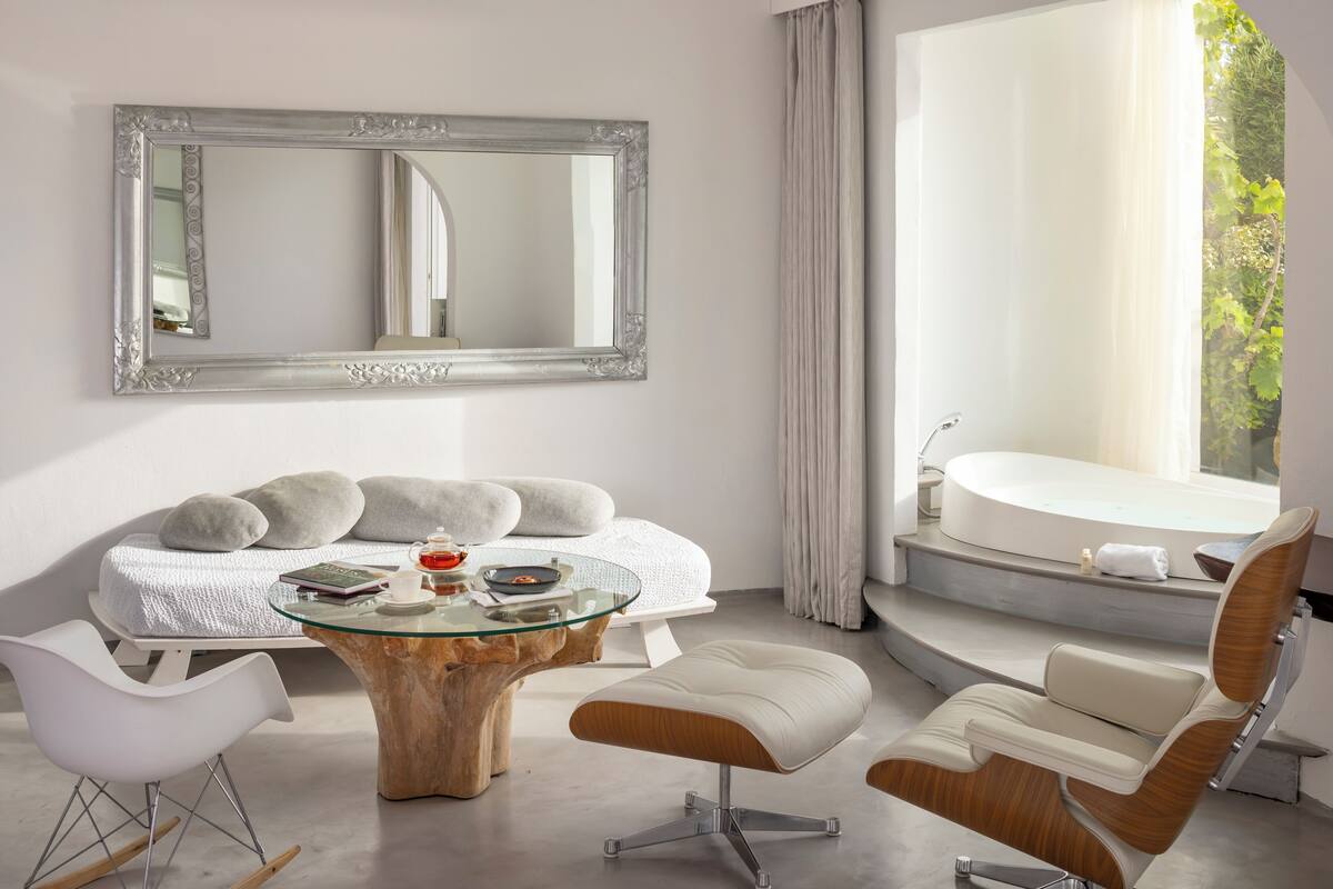 Ein modernes Schlafzimmer mit Glastisch, großem Spiegel, Bett mit weißer Bettwäsche, Sitzecke mit Stühlen und Blick auf eine Badewanne und Grünflächen draußen.
