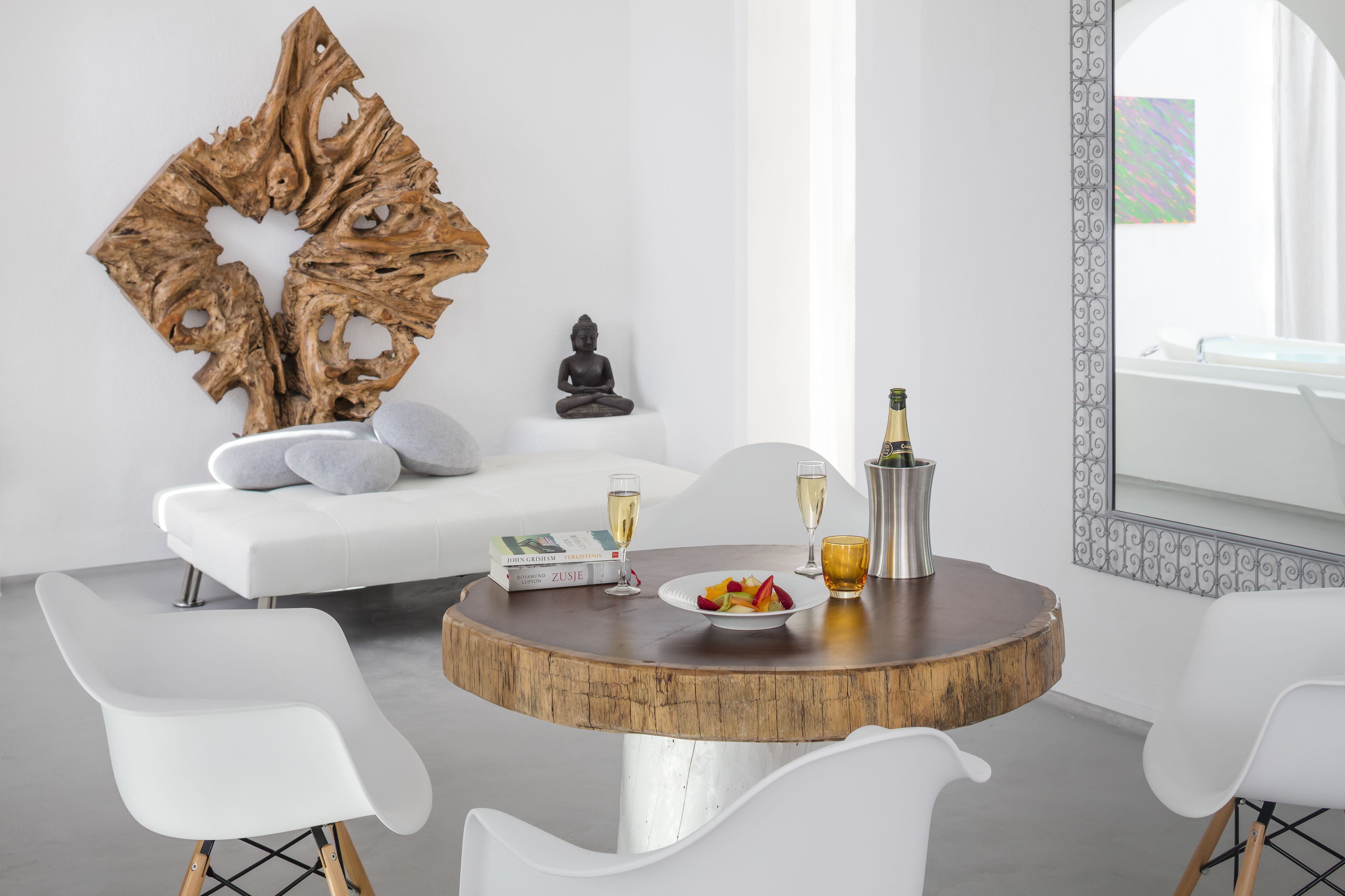 A modern living room with a round wooden table, white chairs, a white sofa, a Buddha statue, and a decorative wooden piece on the wall.