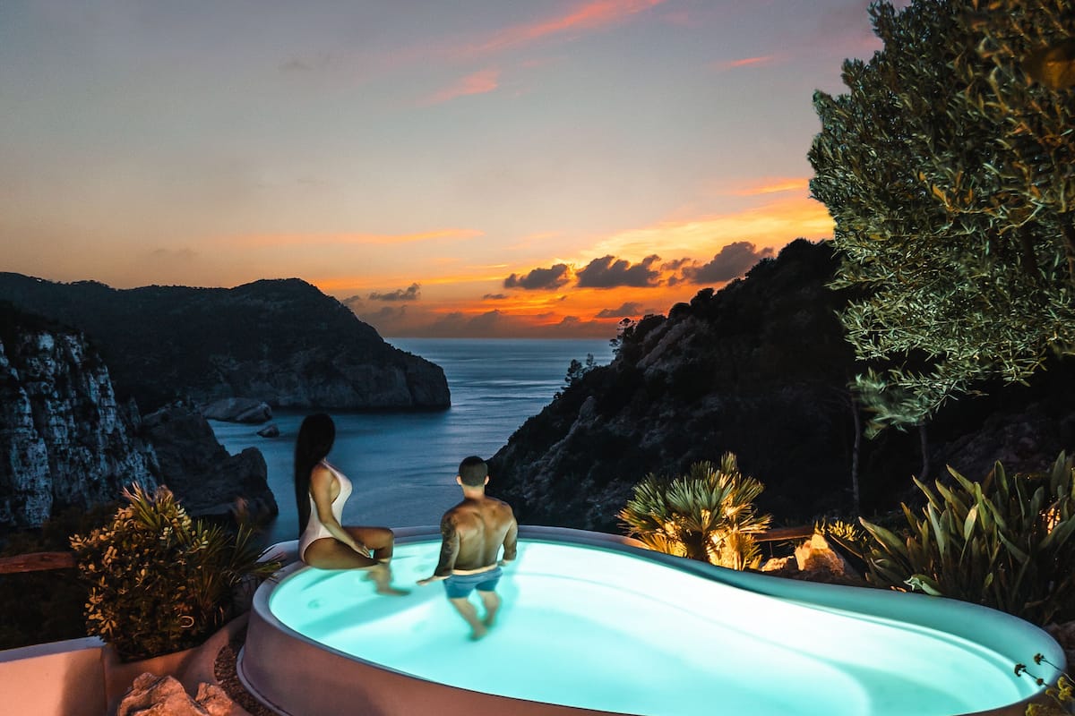 Ein Schwimmbad mit Blick auf den Ozean bei Sonnenuntergang, umgeben von Felsen und Pflanzen.