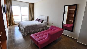 Habitación de hotel con cama, sofá rosa, espejo grande y puerta corrediza de vidrio al balcón.