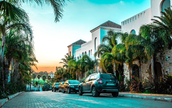 Una calle pavimentada flanqueada por coches aparcados y palmeras, que conduce a un edificio con ventanas arqueadas y tejado de azulejos.