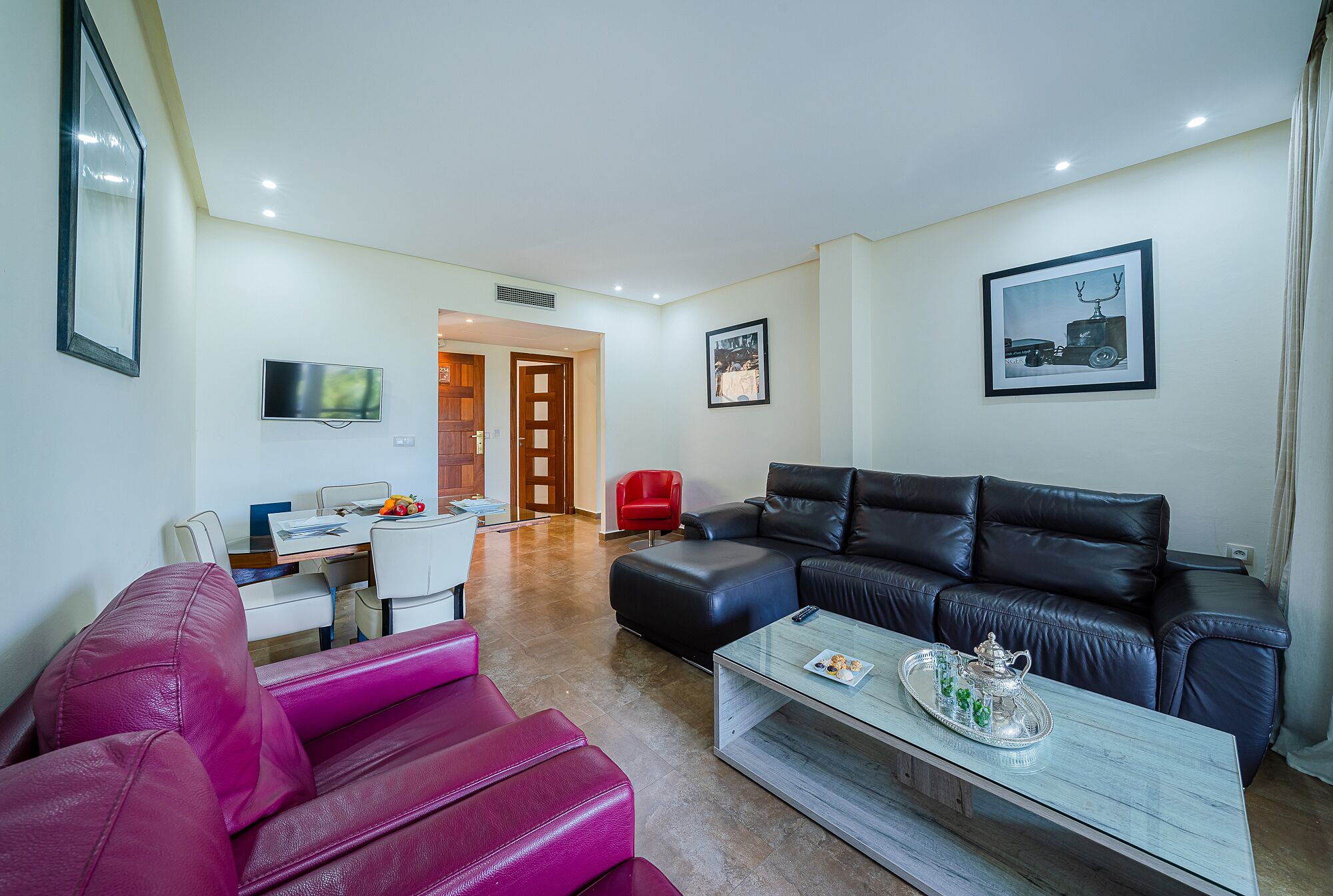A modern living room with a black leather sofa, a red chair, a glass coffee table with a tray, and a TV mounted on the wall.