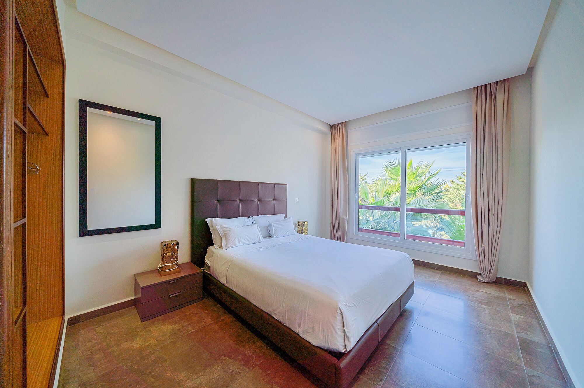 Una camera d'albergo con un letto grande, un comodino, uno specchio e una finestra vista palme.