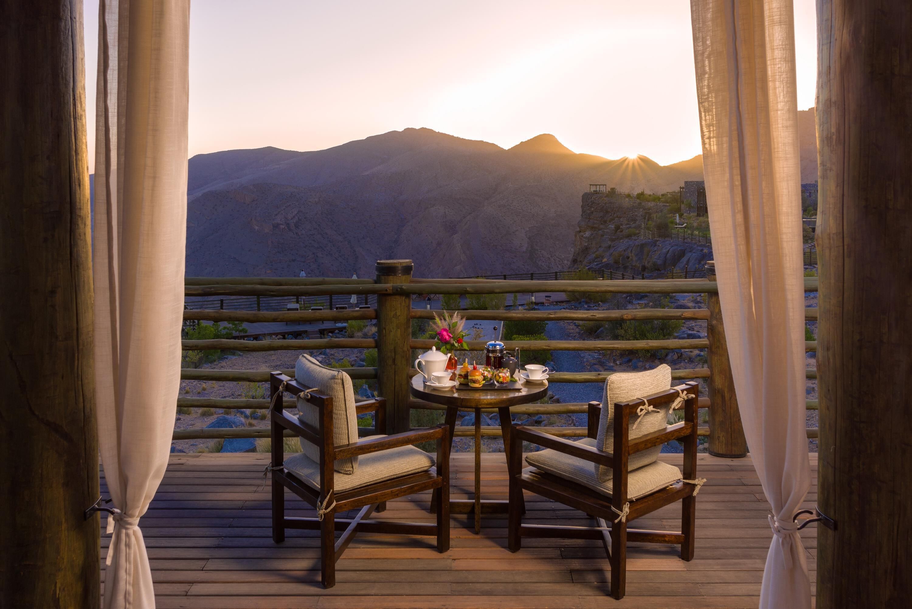 Eine Holzterrasse mit zwei Stühlen und einem für Tee gedeckten Tisch, mit Blick auf eine gebirgige Landschaft.