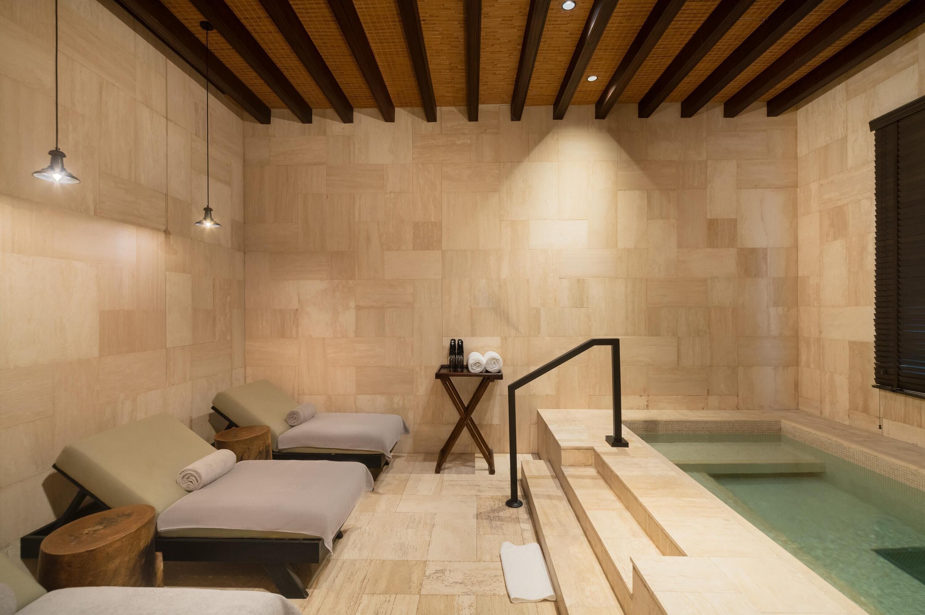 A spa room with wooden flooring, two massage beds, a small table with towels and bottles, and a pool area.