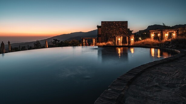 Una piscina infinita tranquila, con borde de piedra, que refleja el cielo vespertino y montañas lejanas.