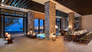 Un elegante comedor interior con pilares de piedra, una piscina y vistas al océano al atardecer.