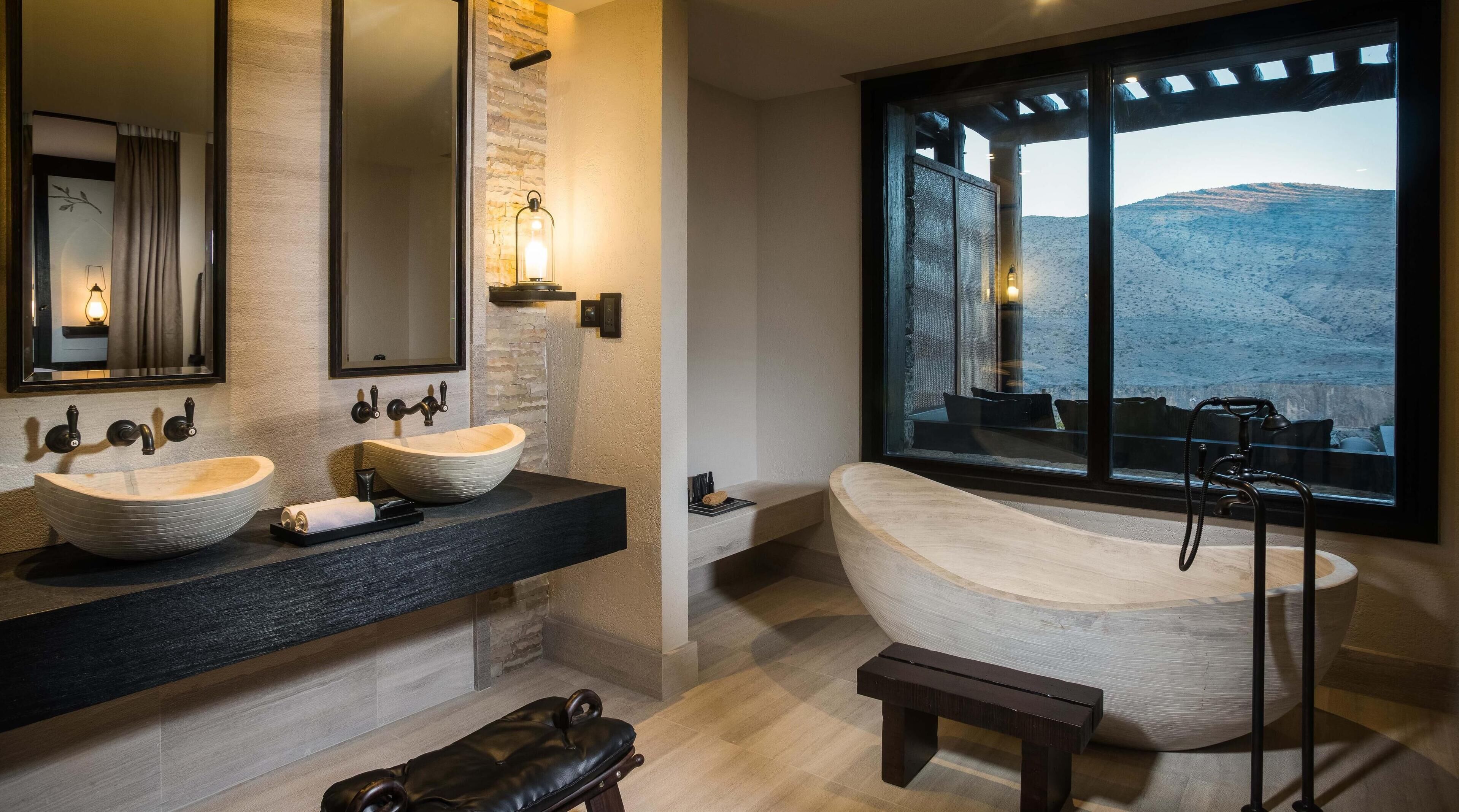 A modern bathroom with two vessel sinks, a freestanding bathtub, and a large window offering a view of a mountainous landscape.