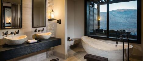 A modern bathroom with two vessel sinks, a freestanding bathtub, and a large window offering a view of a mountainous landscape.