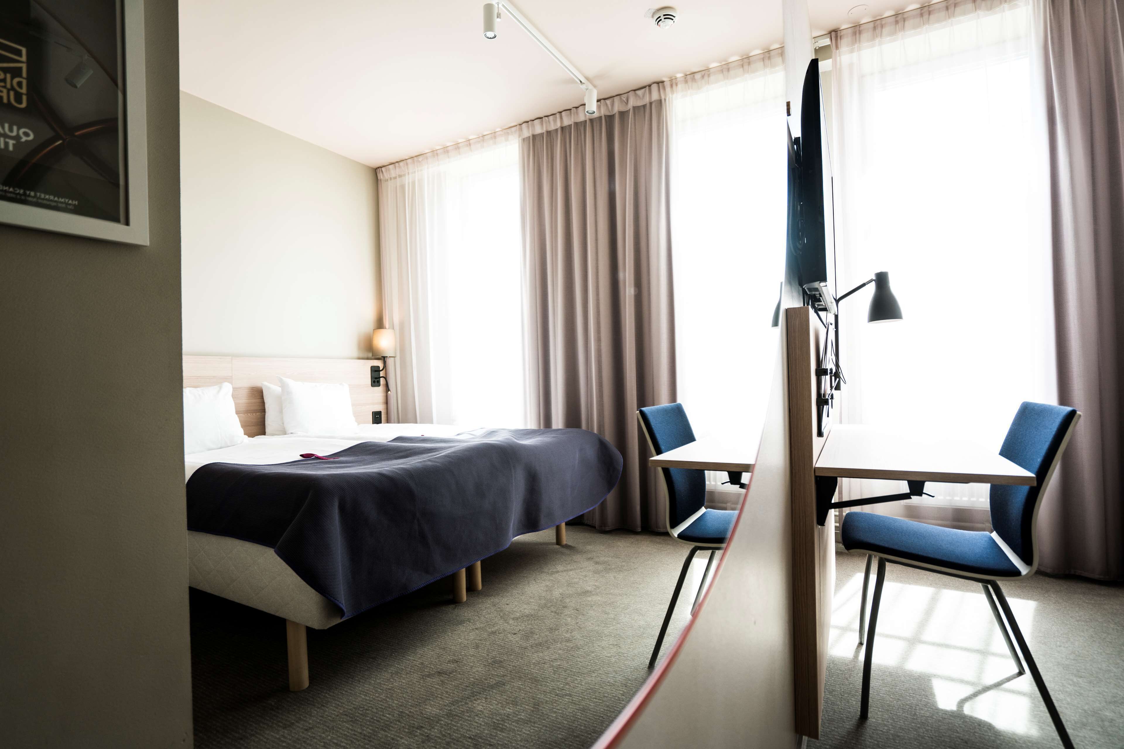 A hotel room with a bed, a desk, two chairs, a lamp, and a TV.