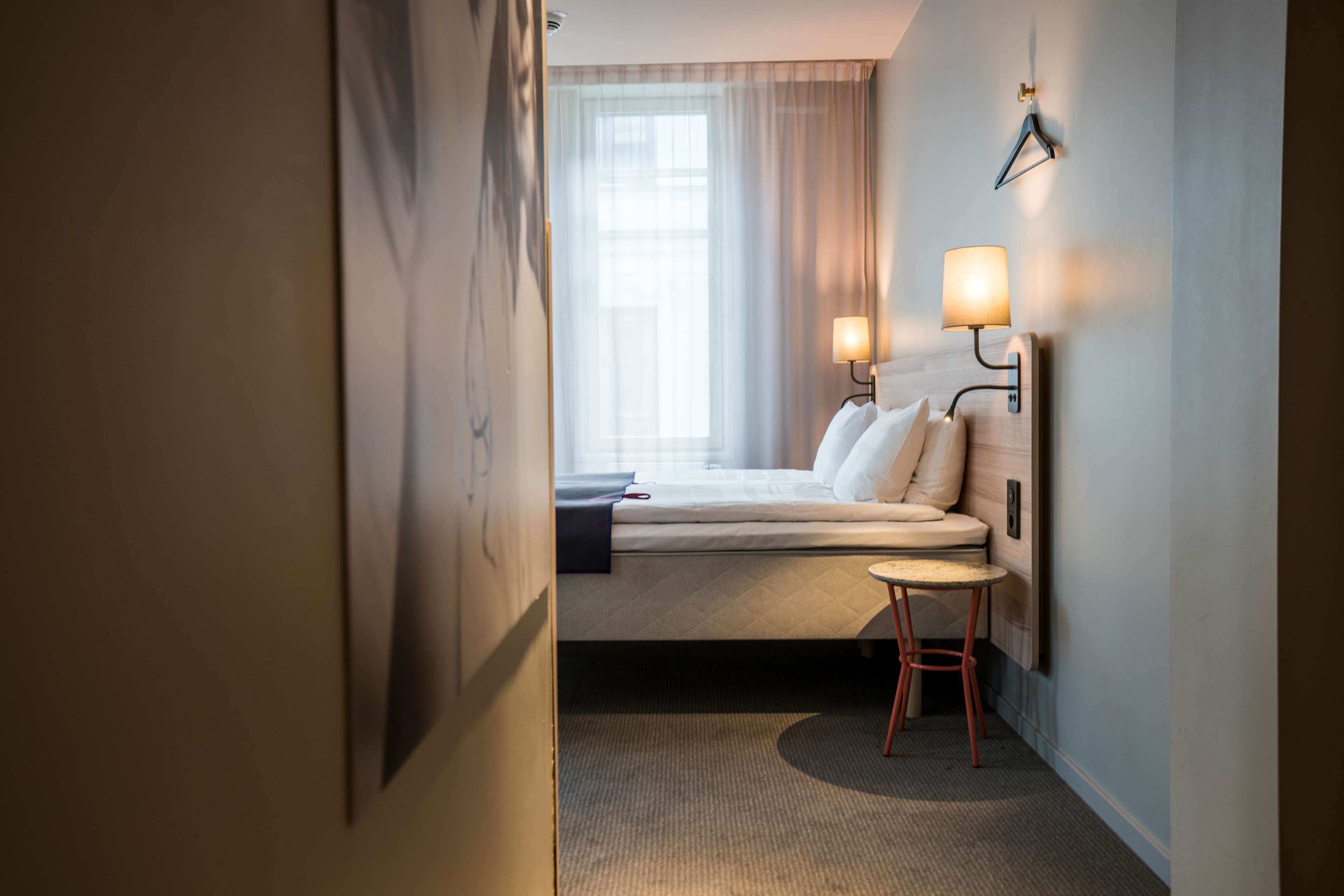 A hotel room with a bed, a small round table, a wall-mounted lamp, and a window with curtains.