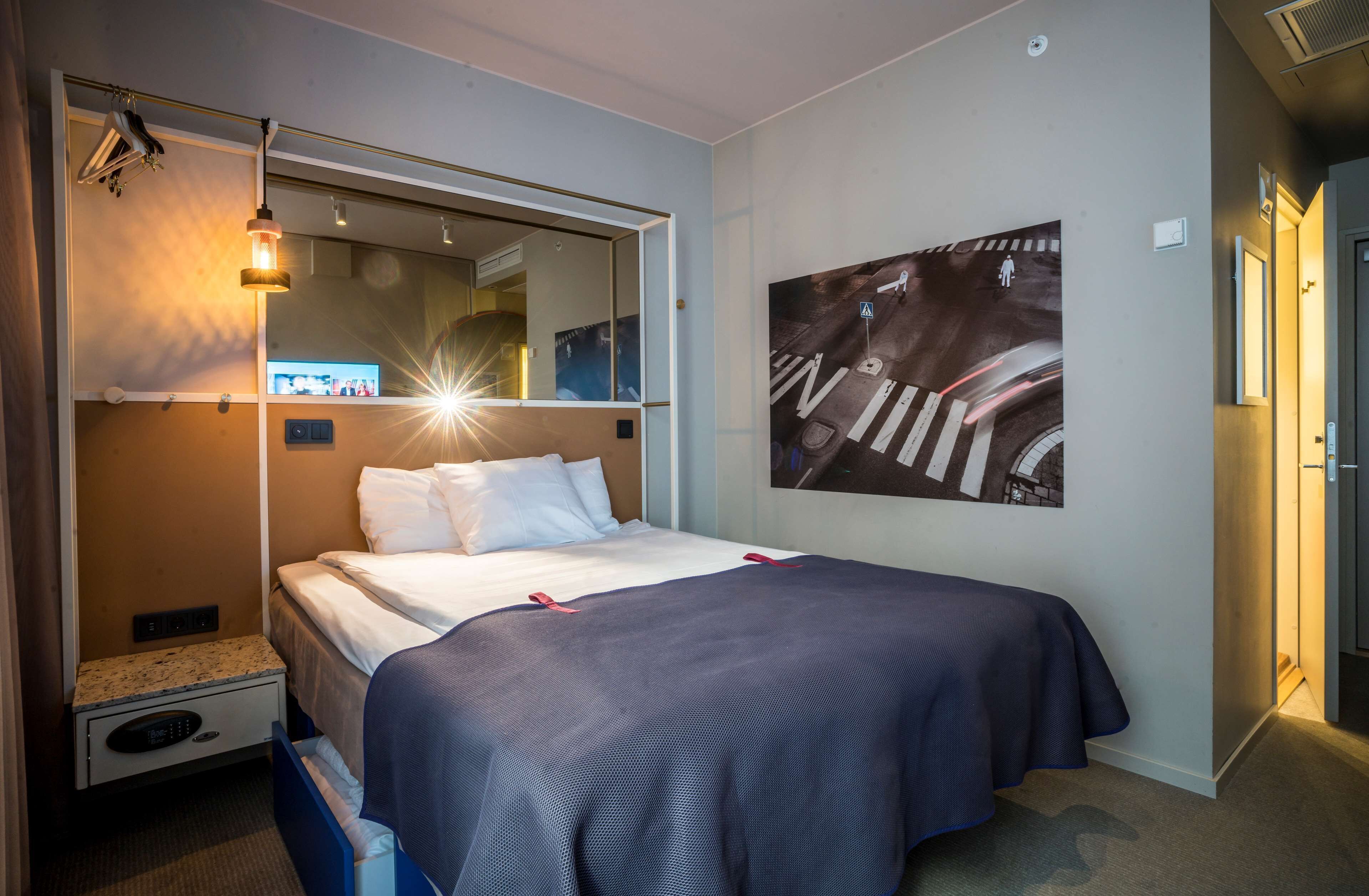 Ett hotellrum med en säng, ett sängbord, en spegel, ett konstverk som hänger på väggen och en taklampa.