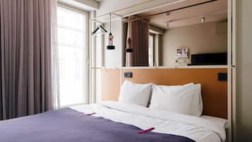 Ett hotellrum med en säng, ett stort fönster med gardiner, en spegel och en hylla med diverse föremål.