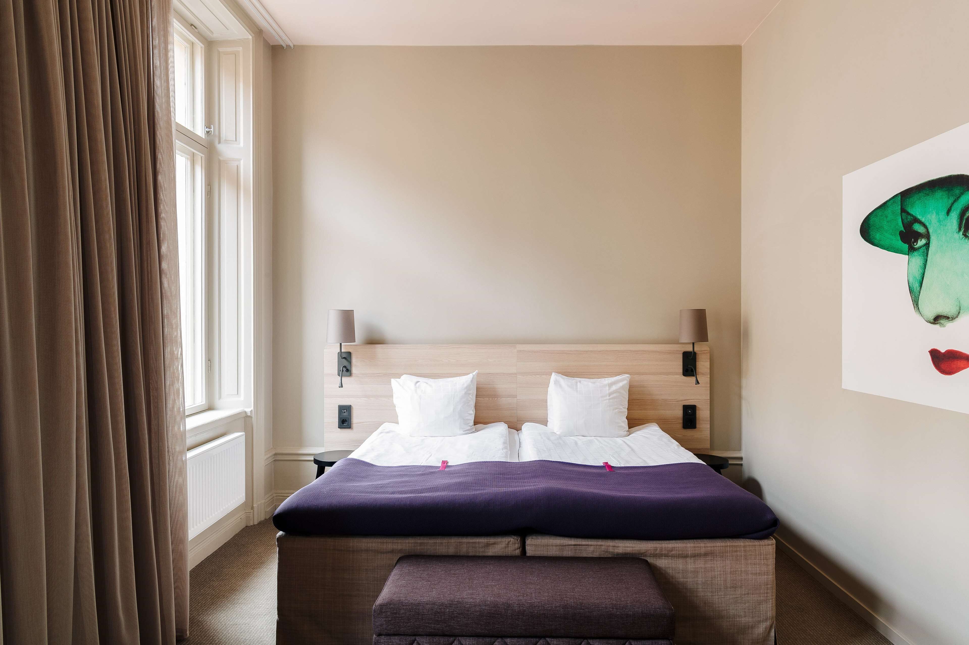 Una habitación de hotel con cama, un banco, una ventana con cortinas, una lámpara de pared y un cuadro.