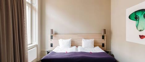 A hotel room with a bed, a bench, a window with curtains, a wall-mounted lamp, and a painting.