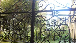 Un cancello in ferro battuto con decorazioni elaborate che conduce a un giardino con alberi e un edificio sullo sfondo.