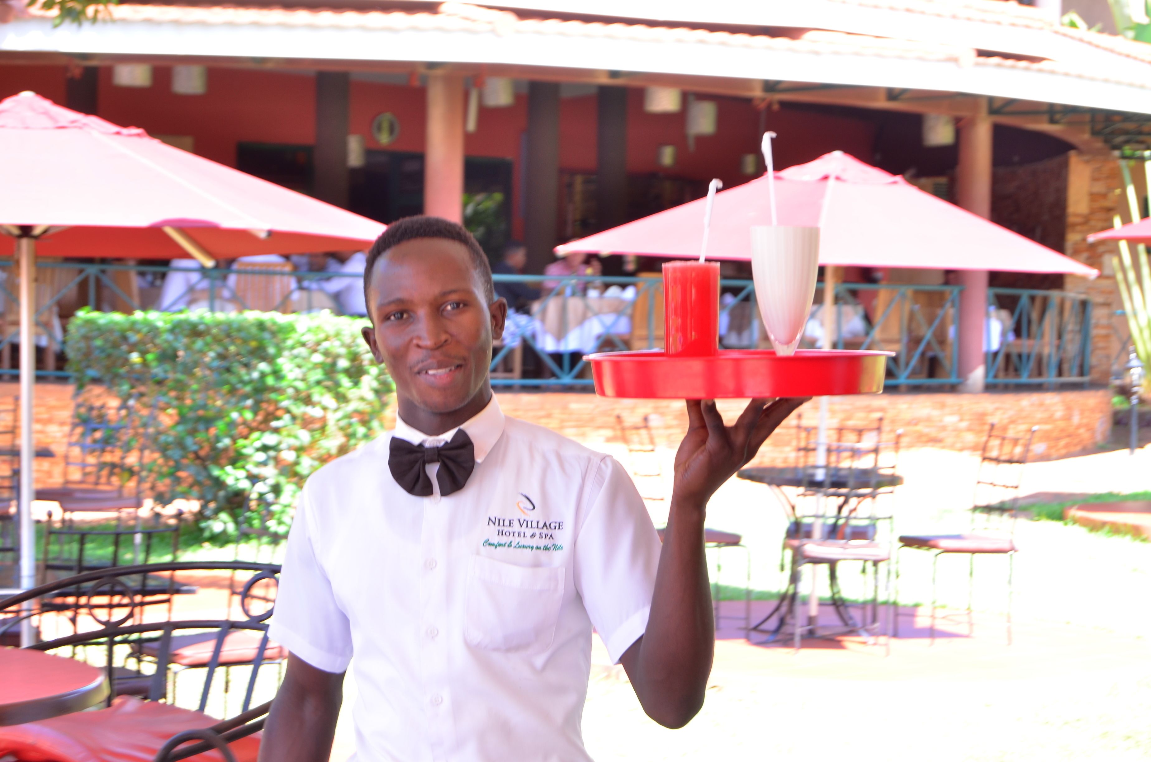 Un membre du personnel de l’hôtel tient un plateau sur lequel se trouvent une boisson et un objet rouge. En arrière-plan, on aperçoit des tables et des chaises à l’extérieur, ainsi que des parasols.