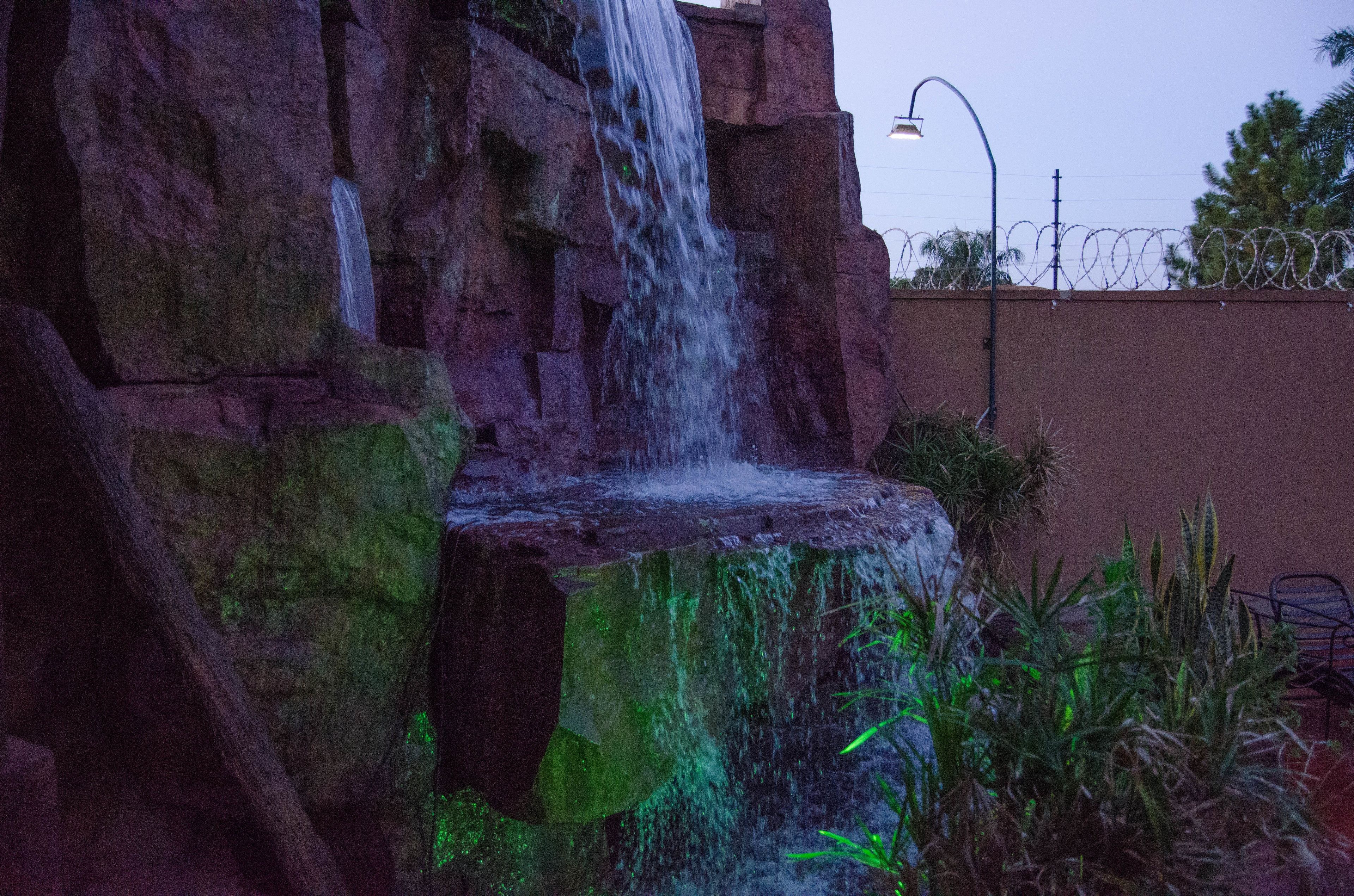 A waterfall cascading down a rocky structure, surrounded by greenery and a barbed wire fence.