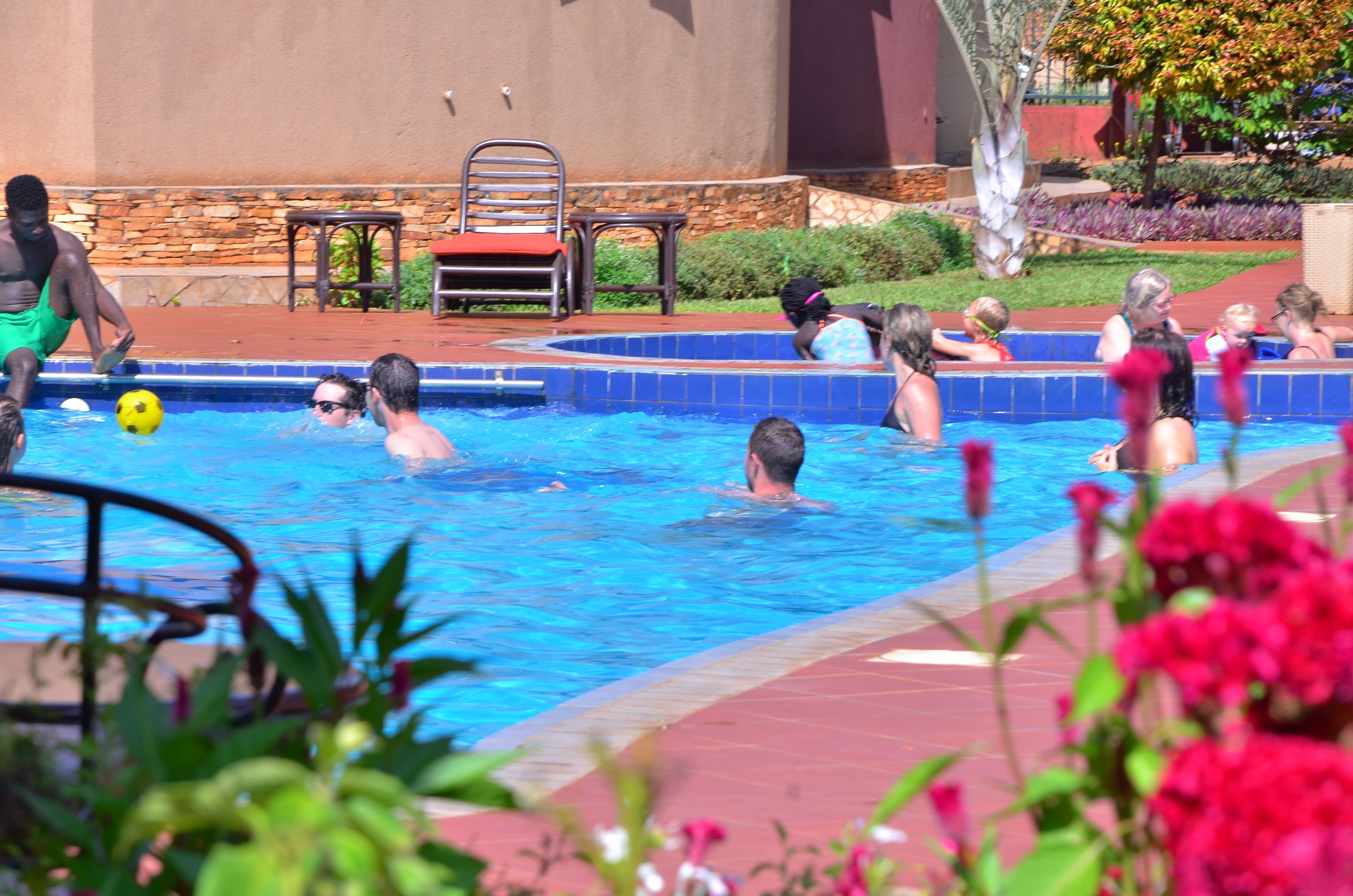 A swimming pool area with people swimming and playing, surrounded by a stone wall and some greenery.