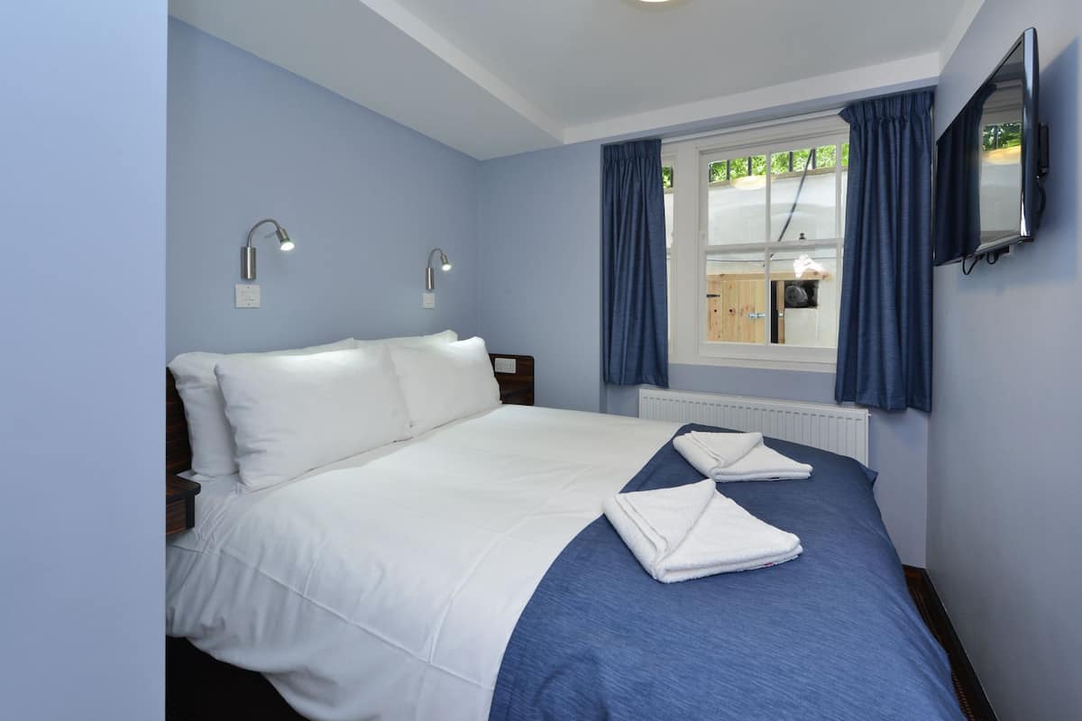 A bedroom with a bed, two bedside lamps, a window with curtains, and a TV mounted on the wall.