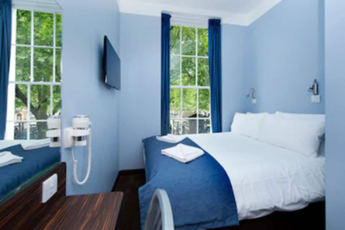 A hotel room with a bed, a bedside table, a TV mounted on the wall, and a window with curtains.