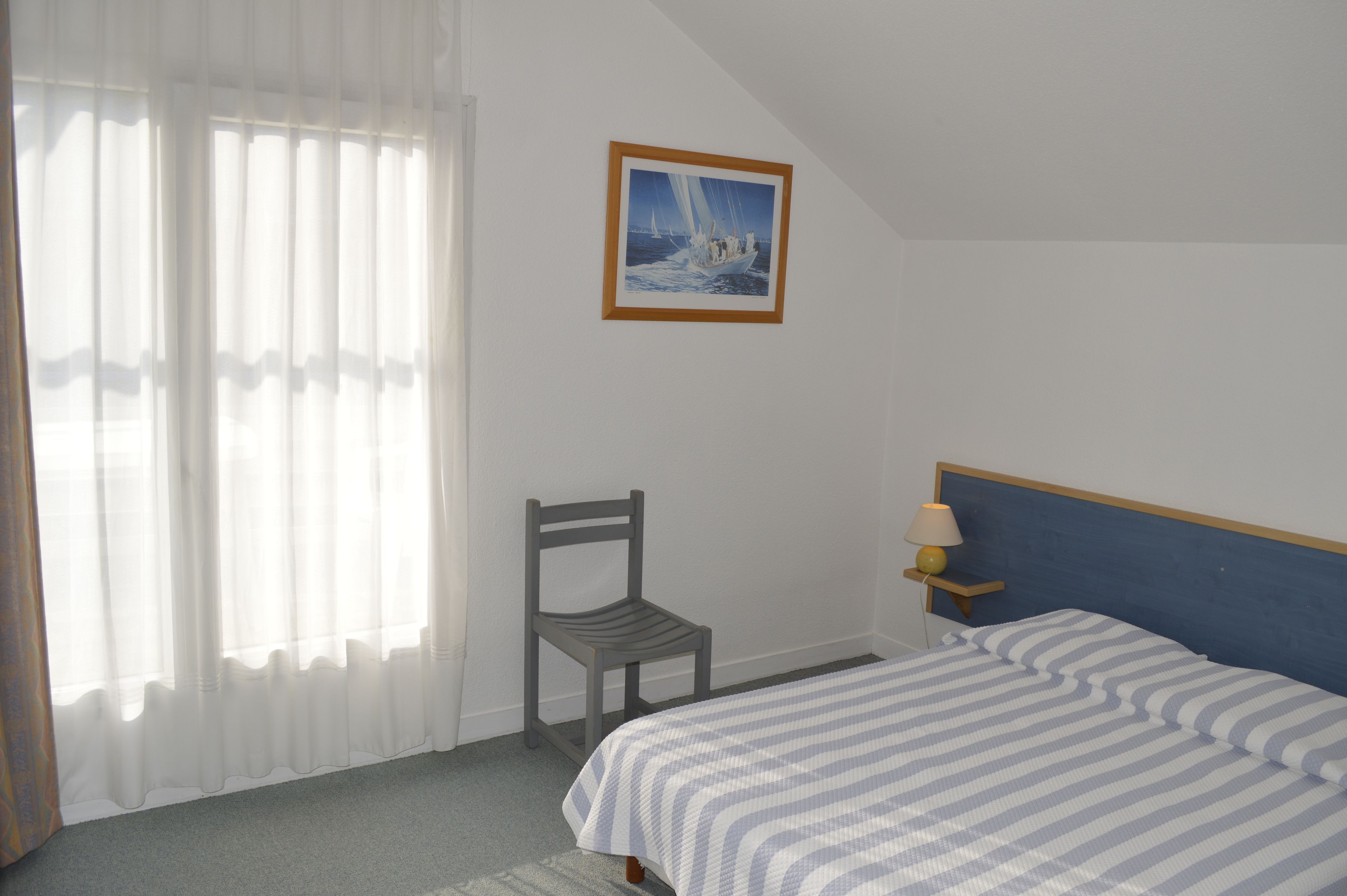 Une chambre à coucher avec un lit, une chaise, une table de chevet avec une lampe, une fenêtre avec des rideaux légers et un tableau encadré au mur.