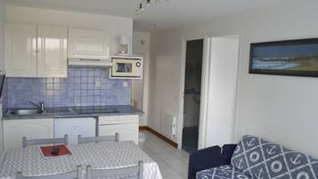 Cocina compacta con armarios blancos, microondas y un lavamanos. Hay una mesa de comedor con sillas y un sofá estampado.