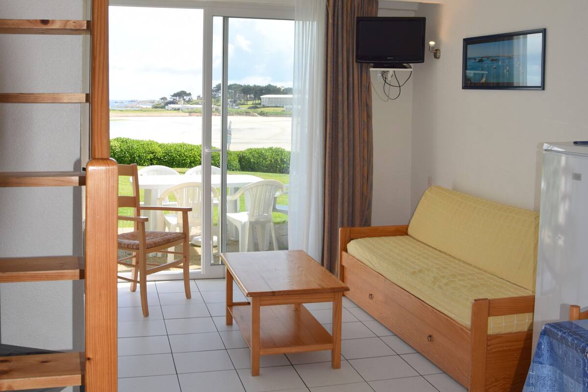 A room with a bed, a TV, a table, and a chair. There is a balcony with a view of the beach.